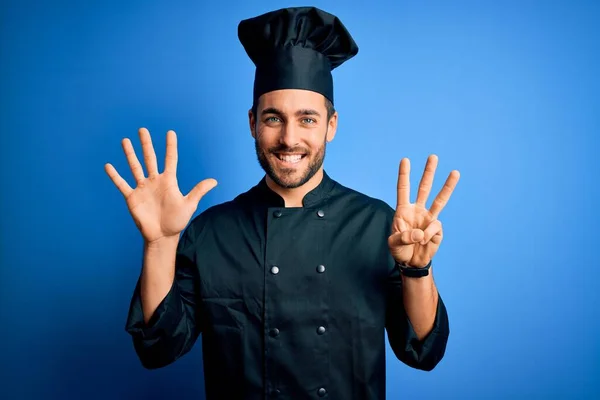 年轻英俊的厨师 留着胡子 身穿炊事服 头戴蓝色背景的帽子 露出八号手指 面带微笑 自信而快乐 — 图库照片