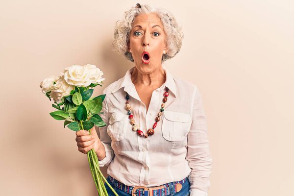 Senior grey-haired woman holding flowers scared and amazed with open mouth for surprise, disbelief face 