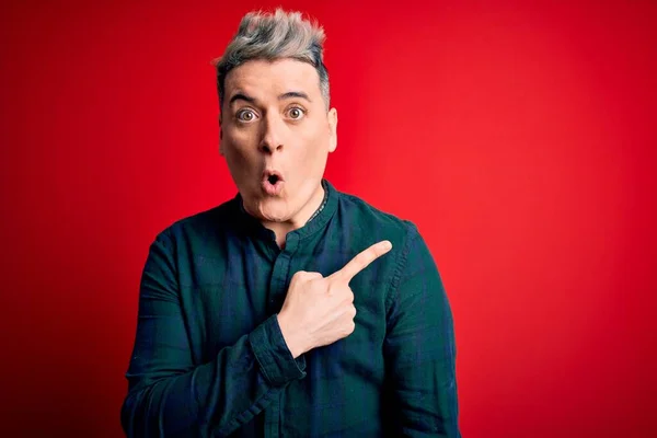 Young handsome modern man wearing elegant green shirt over red isolated background Surprised pointing with finger to the side, open mouth amazed expression.