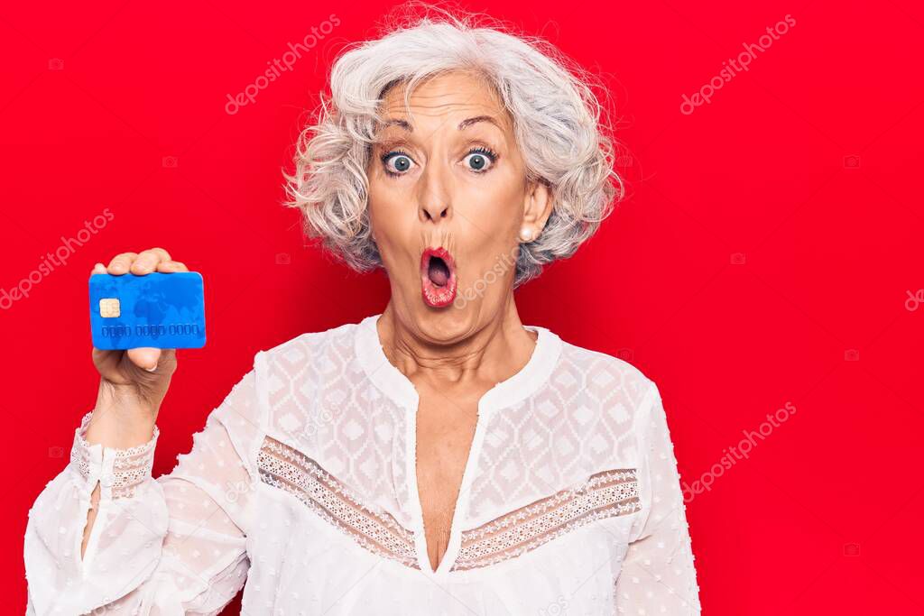 Senior grey-haired woman holding credit card scared and amazed with open mouth for surprise, disbelief face 
