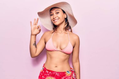 Bikini giyen, şapkalı, olumlu gülümseyen genç bir kadın el ve parmaklarıyla imza atıyor. başarılı ifade. 