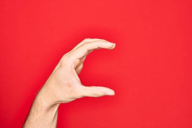 Kafkasyalı genç adam parmaklarını izole edilmiş kırmızı arka plan üzerinde gösteriyor ve görünmez şeyi alıyor, parmakları boşluk gösteren nesneleri tutuyor.