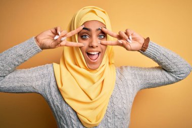 Genç ve güzel Afro-Amerikalı kız izole edilmiş sarı arka plan üzerine Müslüman başörtüsü takıyor. Yüzünde parmaklarıyla barış sembolü yapıyor, neşeyle gülümsüyor zaferini gösteriyor.