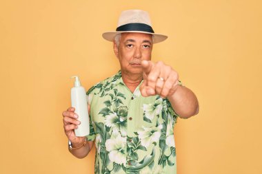 Orta yaşlı, kır saçlı, yaz şapkası takan, elinde güneş kremi tutan, kamerayı işaret eden, el işareti olan, ön taraftan olumlu ve kendinden emin bir jest yapan yaşlı bir adam.