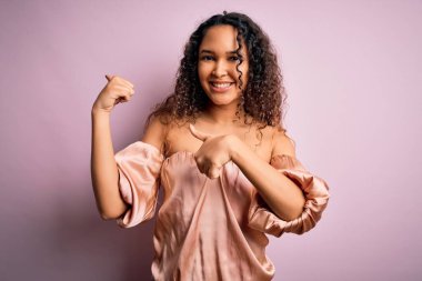 Kıvırcık saçlı, rahat tişörtlü, pembe arka planda duran, eli ve başparmağı havada, kendine güvenen genç ve güzel bir kadın.