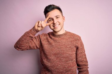 Pembe izole edilmiş arka plan üzerine sıradan bir kazak giyen yakışıklı beyaz adam barış sembolü yapıyor. Yüzünde parmaklar, gülümseyerek zaferi gösteriyor.