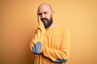 Sarı arka planda dikilen, sakallı yakışıklı kel adam kollarını kavuşturup depresyondan sıkılmış ve yorgun görünüyor..