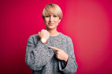 Modern kısa saçlı, pembe arka planda günlük süveter giyen genç sarışın kadın saati, sabırsızlığı ve rahat bir ifadeyle kameraya bakıyor.