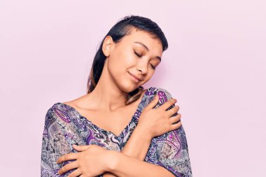 Gündelik kıyafetler giyen, mutlu ve olumlu bir şekilde kendine sarılan, kendinden emin gülümseyen genç bir kadın. Sevgi ve özgüven. 