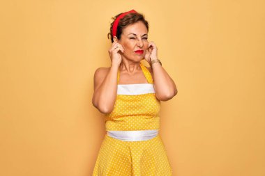 Orta yaşlı bir kadın, 50 'li yaşlarda, sarı arka plan üzerine eski tarz bir elbise giyip yüksek sesli müzik sesi için parmaklarıyla kulaklarını kapatıyor. Sağır kavramı.