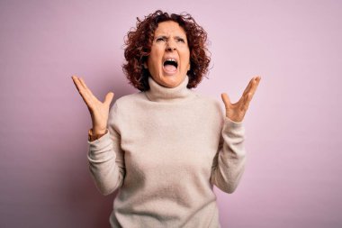 Orta yaşlı güzel, kıvırcık saçlı kadın pembe arka plan üzerine balıkçı yaka kazak giyiyor deli gibi bağırıyor ve agresif bir ifade ve kolla bağırıyor. Hayal kırıklığı konsepti.