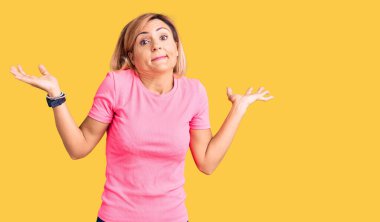 Young blonde woman wearing sportswear clueless and confused expression with arms and hands raised. doubt concept.  clipart