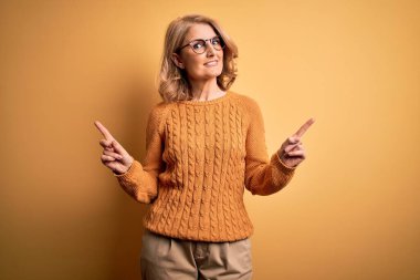 Orta yaşlı güzel sarışın kadın sarı arka planda rahat süveter ve gözlük takıyor. Kendine güvenen, farklı yönleri gösteren bir gülümseme. Reklam için boşluk kopyala