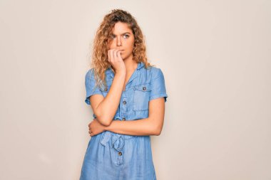 Mavi gözlü genç bir kadın beyaz arka planda rahat bir kot elbise giyiyor gergin ve elleri ağızdan tırnak yerken gergin görünüyor. Endişe sorunu.