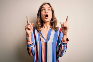 Renkli çizgili tişörtlü genç sarışın kız izole arka planda şaşkın şaşkın yukarı bakıyor ve parmaklarıyla işaret ediyor ve kollarını kaldırıyor..