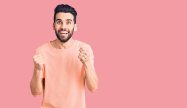 Sakallı, sıradan tişörtlü genç ve yakışıklı bir adam kolları havada ve gözleri açık bir şekilde başarıyı kutluyor. Kazanan konsept. 