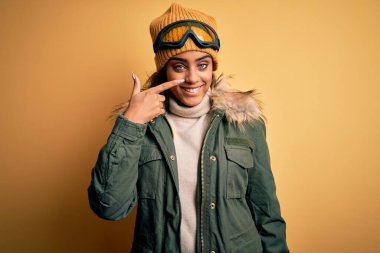 Genç Afro-Amerikan kayakçı kız kar spor giyim ve kayak gözlüğü takıyor. El ve burunla işaret ediyor. Neşeli gülümsüyor. Güzellik konsepti