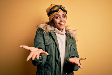 Genç Afrikalı Afro-Amerikan kayakçı kız kar sporu giysisi ve kayak gözlüğü takıyor. Kolları açık gülümsüyor. Dostça karşılama, olumlu ve kendinden emin selamlar.