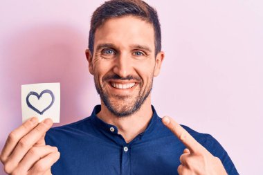Pembe arka planda kalp şeklinde bir hatıra kâğıdı tutan genç, yakışıklı adam mutlu bir şekilde eli ve parmağı ile işaret ediyor.