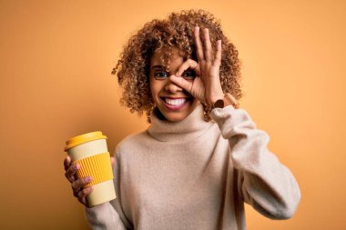 Genç Afro-Amerikan kıvırcık kadın, sarı arka planda bir fincan kahve içiyor, gülümsüyor, el ele imzasını atıyor, parmaklarının arasından bakıyor.