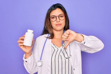 Mor arka planda ilaçlarını tutan genç profesyonel doktor kadın. Kızgın yüzlü, negatif işaretli, baş parmakları aşağıda, reddedilme kavramından hoşlanmadığını gösteren.
