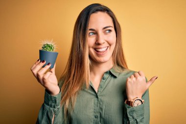 Mavi gözlü, genç, sarışın bir kadın, elinde küçük bir kaktüs tenceresi, sarı halkalı yeri işaret ediyor ve gülümseyen bir yüzle baş parmağıyla onu gösteriyor.