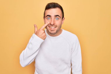 Mavi gözlü, sarı arka planda dikilen, el parmağı ve burunla işaret eden, neşeli gülümseyen, sıradan bir süveter giyen genç yakışıklı adam. Güzellik konsepti