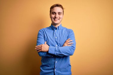 İzole edilmiş sarı arka plan üzerinde sıradan çizgili tişört giyen genç, kızıl saçlı adam kameraya bakarak kollarını kavuşturarak gülümsüyor. Pozitif kişi.