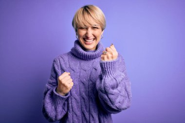 Kısa saçlı, mor arka planda balıkçı yaka kazak giyen genç sarışın kadın, kolları havada ve gözleri kapalı olarak başarıyı kutluyor ve şaşırıyor. Kazanan konsept.