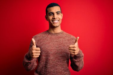 Genç, Afro-Amerikan bir adam, günlük süveter giyip kırmızı arka plan burcunun üzerinde duruyor. Elleriyle olumlu jest yapıyor, gülümsüyor ve mutlu. Neşeli ifade ve kazanan jest.