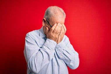 Orta yaşlı, yakışıklı, beyaz bir adam, kırmızı arka plan üzerinde gözlüklü, normal çizgili tişört giyiyor ve ağlarken suratını ellerle kapatıyor. Depresyon kavramı.