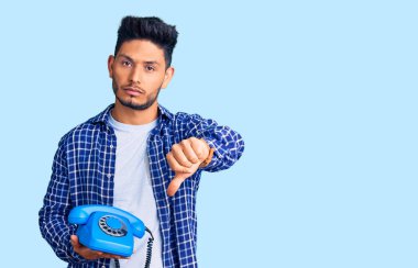 Yakışıklı Latin Amerikalı genç adam elinde kızgın yüzlü, antika bir telefon tutuyor. Eksi işareti baş parmakları aşağıda, reddedilme konsepti ise reddedilmiş. 