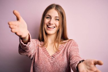 Genç, güzel, kızıl saçlı bir kadın izole edilmiş pembe arka plan üzerine günlük süveter giyerek kameraya bakıyor ve sarılmak için kollarını açmış gülümsüyor. Mutluluğu kucaklayan neşeli bir ifade..