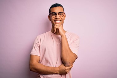 Yakışıklı, Afro-Amerikan bir adam pembe arka plan üzerinde günlük tişört ve gözlük takıyor. Kameraya kollarını kavuşturup, ellerini çenesine kaldırarak gülümsüyor. Olumlu düşünüyorum..