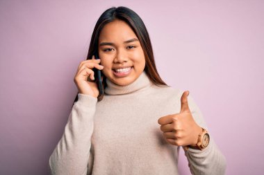 Genç Asyalı kız akıllı telefondan konuşuyor pembe arka planda büyük bir gülümseme ile konuşuyor tamam işareti yapıyor, parmaklarıyla başparmağını kaldırıyor, mükemmel bir işaret