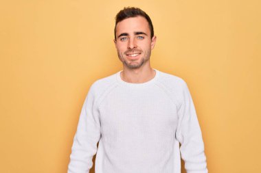 Mavi gözlü, sarı arka planda dikilen, mutlu ve havalı gülümseyen sıradan bir süveter giyen genç yakışıklı bir adam. Şanslı kişi..