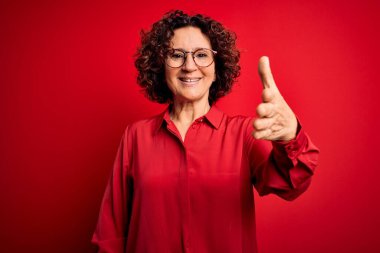 Orta yaşlı güzel kıvırcık saçlı kadın kırmızı arka planda günlük gömlek ve gözlük takıyor dostça gülümseyerek selamlama ve hoş geldin diyor. Başarılı bir iş..