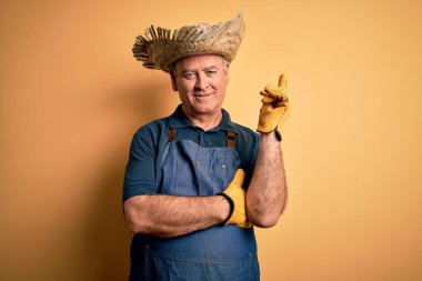 Orta yaşlı, beyaz önlüklü ve şapkalı, izole edilmiş sarı arka planda, yüzünde kocaman bir gülümseme, eli ve parmağı kameraya bakan bir çiftçi..
