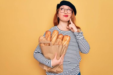 Bere takan genç kızıl saçlı bir kadın sarı arka planda ekmek taşıyan kağıt bir çanta taşıyor. Ciddi bir yüz. Soru sormayı düşünüyor, kafası karışık bir fikir.