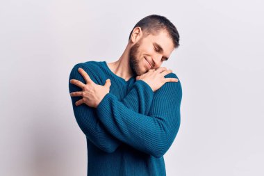 Genç yakışıklı bir adam sıradan bir süveter giyip kendine sarılıyor mutlu ve pozitif, kendinden emin gülümsüyor. Sevgi ve özgüven. 