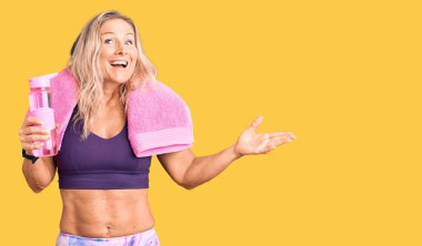 Orta yaşlı, sarışın, spor giysili, havluyla gezen ve elinde su şişesi olan bir kadın zaferi mutlu bir gülümsemeyle kutluyor. 