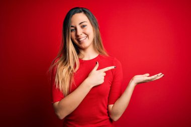 Kırmızı arka planda sıradan tişörtler giyen mavi gözlü genç ve güzel sarışın kadın kameraya hayret ediyor ve gülümsüyor..