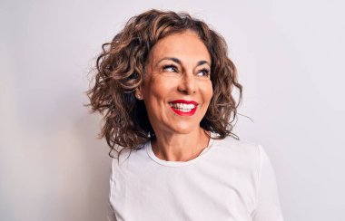 Orta yaşlı, güzel, esmer bir kadın beyaz arka planda dikilmiş rahat bir görünüm ve kendine güvenen bir gülümseme takınıyor..