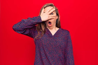 Genç, güzel, sarışın bir kadın. Kırmızı arka planda dikiliyor. Şok olmuş bir şekilde yüzünü ve gözlerini kapatıyor. 