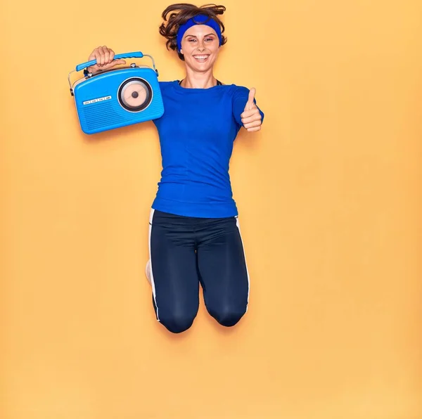 若い美しいスポーツ女性の音楽を聞いて幸せな笑顔 ジャンプとともに笑顔上の顔を保持ヴィンテージラジオDoing Signとともに親指アップ上の隔離された黄色の背景 — ストック写真