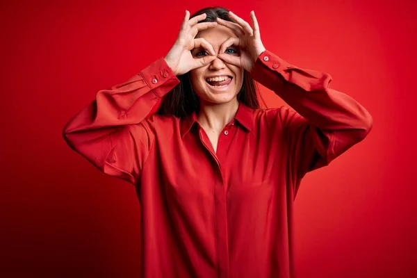 Young beautiful woman with blue eyes wearing casual shirt standing over red background doing ok gesture like binoculars sticking tongue out, eyes looking through fingers. Crazy expression.