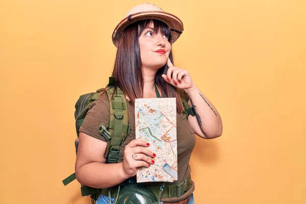 Young plus size woman wearing explorer hat holding map serious face thinking about question with hand on chin, thoughtful about confusing idea