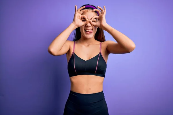 年轻漂亮的女运动员穿着运动服 穿着紫色背景的运动服 做着像双筒望远镜一样的手势 伸出舌头 眼睛透过手指看 疯狂的表情 — 图库照片