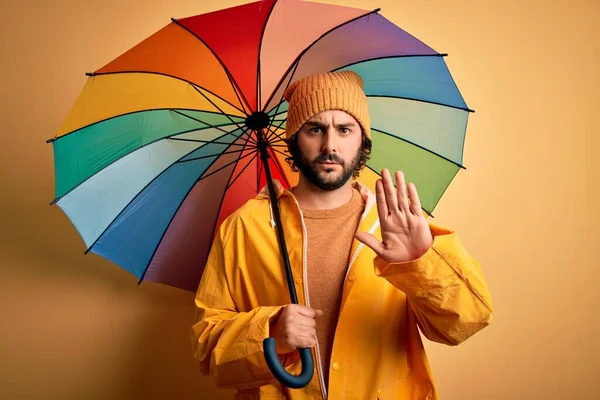 年轻英俊的大胡子男子 雨衣雨天身穿雨衣 手持五彩缤纷的雨伞 张开双手 严肃而自信地做停止手势 做防守动作 — 图库照片