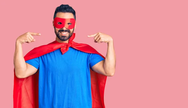 Young hispanic man wearing super hero costume looking confident with smile on face, pointing oneself with fingers proud and happy.